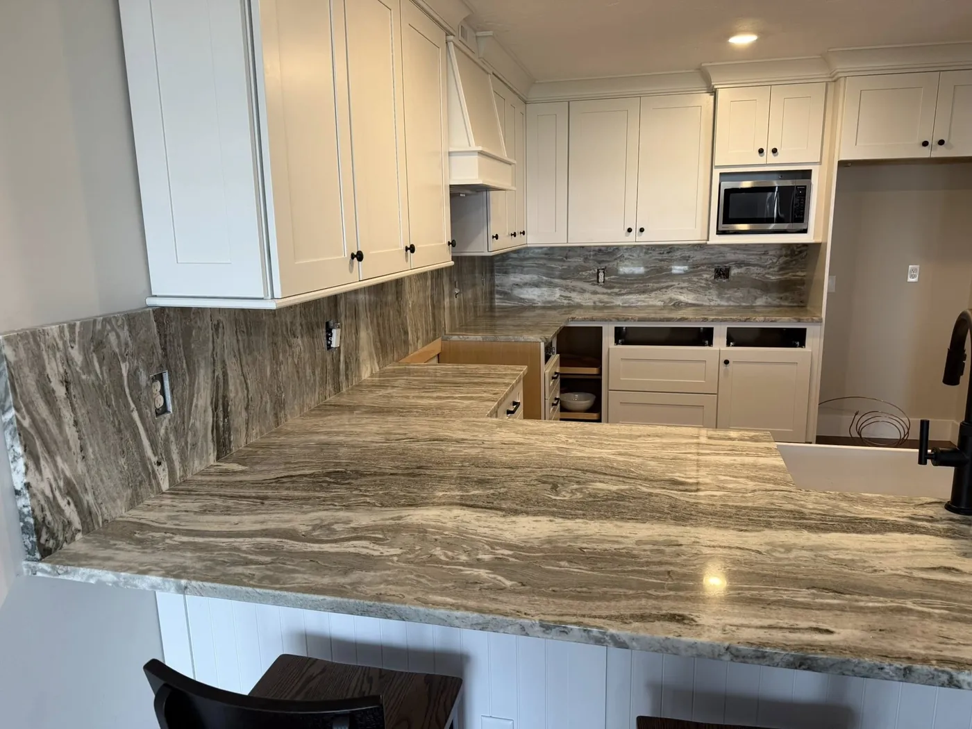 Flowing gray and cream stone repeated across kitchen countertops and a full-height backsplash beneath white cabinets