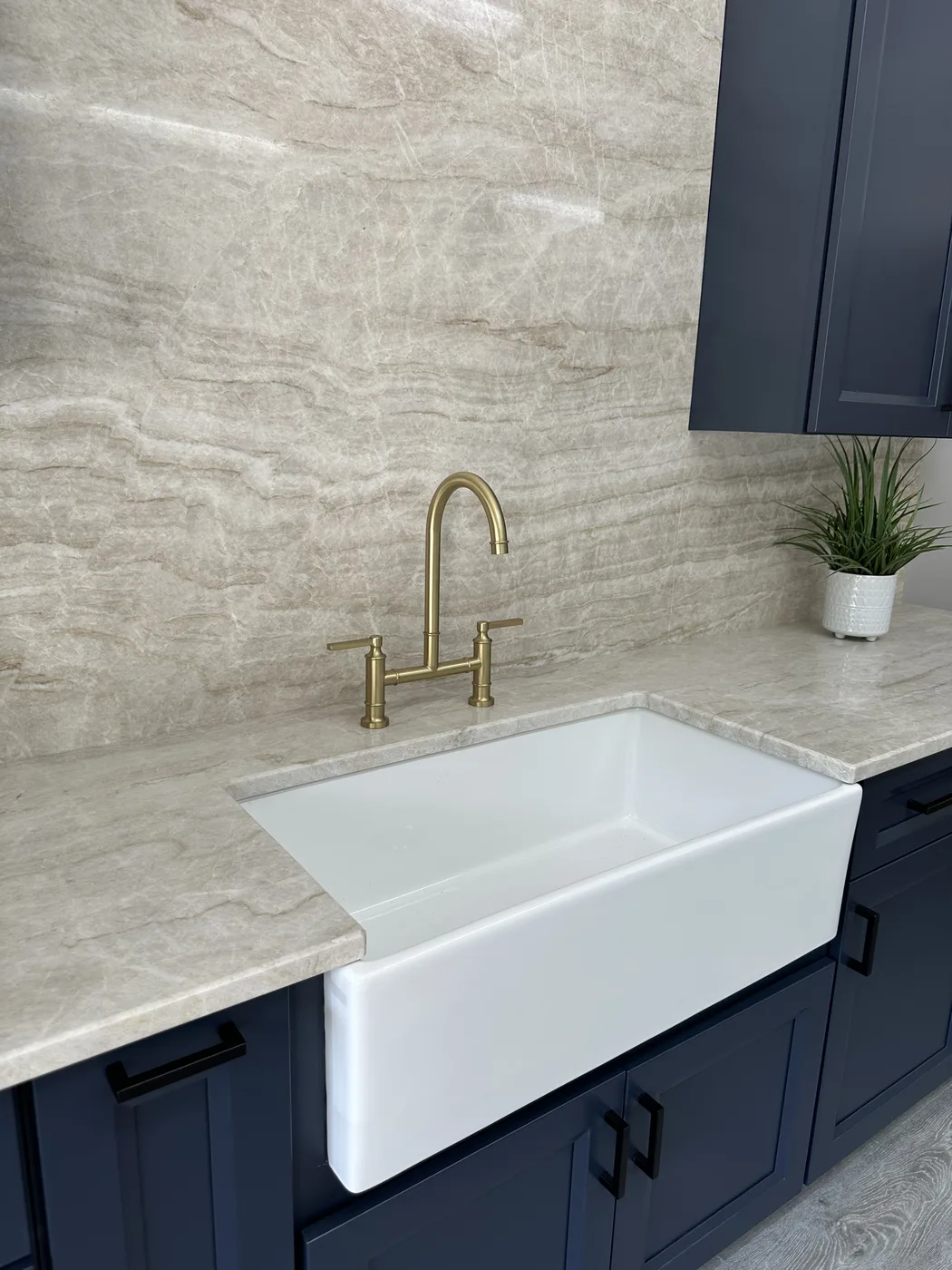 White farmhouse sink surrounded by cream stone countertops and a full-height stone backsplash with a brass faucet and dark blue cabinets
